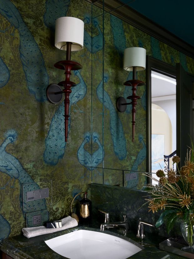 A dramatic, richly decorated bathroom vanity featuring a white inset basin set into a dark stone benchtop, polished chrome tapware, mirrored cabinetry or wall panels, and distinctive wall-mounted lamps with deep red decorative stems. The walls are covered in a vivid blue-green peacock-patterned decorative surface, creating a luxurious, eclectic atmosphere. A tall window or opening brings in daylight and reveals an exterior view.