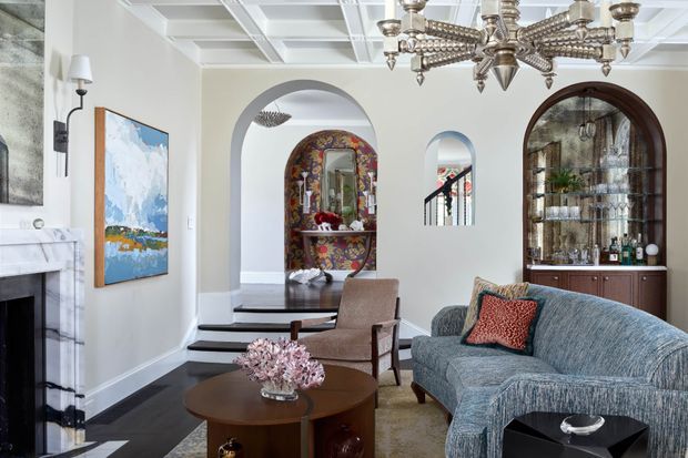 Elegant traditional living room with a coffered ceiling, ornate chandelier, marble fireplace, arched openings, dark timber flooring, and a curved blue sofa. A built-in arched glass-front drinks cabinet and richly patterned decorative alcove add refined character, while a dining or stair area is visible through the openings.