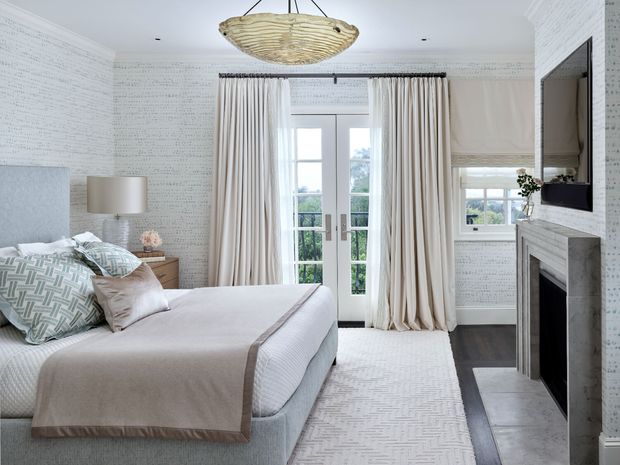 Bright, refined bedroom with a large upholstered bed, layered neutral bedding, patterned pillows, a pale area rug, and full-height cream curtains framing glazed French doors. The room features white patterned wallpaper, a marble or stone fireplace surround, dark timber flooring, a light timber bedside table, and a sculptural woven pendant light. A window seat or low upholstered bench is visible beside the fireplace, with greenery seen outside through the glazing.