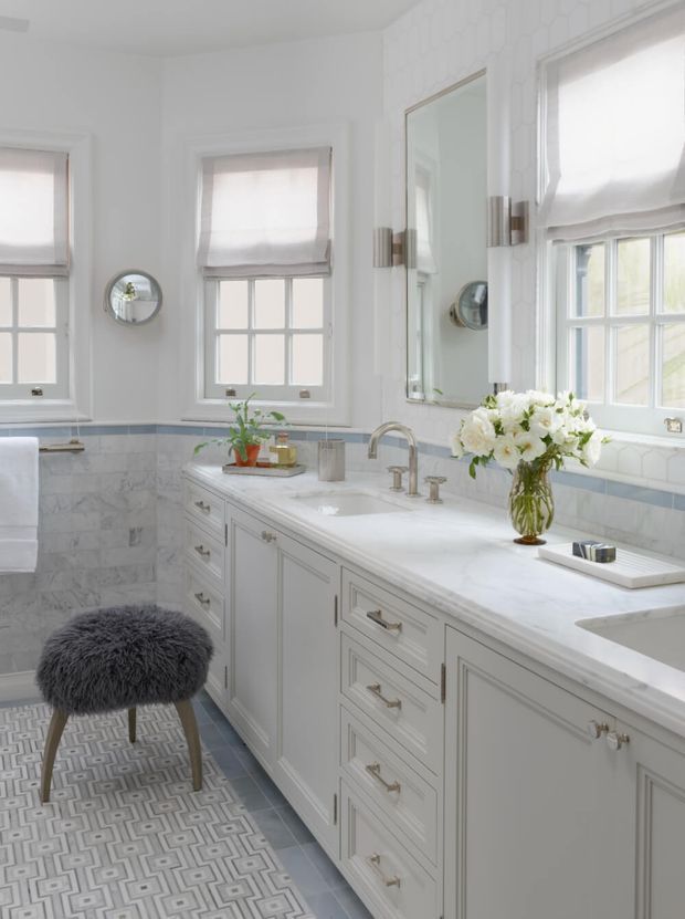 Bright, refined bathroom with a long double-sink vanity, pale cabinetry, white marble-look benchtop, marble wall surfaces, patterned floor tile, and polished metal tapware. Multiple windows with soft Roman blinds provide abundant natural light. A large wall mirror, round magnifying mirror, decorative sconces, and a furry stool complete the elegant traditional interior.