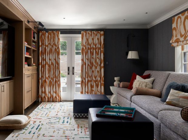 A richly layered living room with dark charcoal walls, white crown moulding, built-in light timber cabinetry and shelving, and a large grey upholstered sofa. Patterned orange-and-cream curtains frame glazed French doors, while a matching Roman blind covers the side window. The room includes a black ottoman coffee table, blue ottoman, patterned rug, floor lamp, cushions, and decorative accessories. The overall look is tailored, comfortable, and colourfully eclectic with traditional architectural detailing.