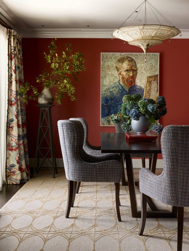 A richly styled dining room with a deep red accent wall, patterned floor-to-ceiling curtains, a dark wood dining table, upholstered dining chairs, and a large ornate pendant light. A framed portrait artwork, sculptural potted plant, floral arrangement, and patterned carpet add layered traditional character. White ceiling mouldings and recessed lights complete the room.