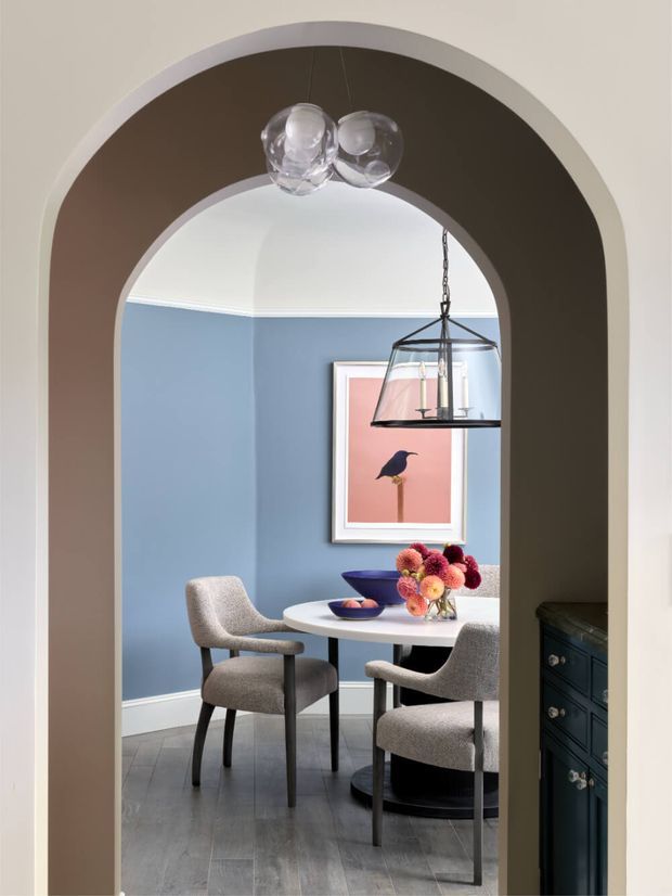A refined dining room viewed through a large rounded archway. The room features muted blue walls with white trim, dark grey timber-look flooring, a round white dining table, upholstered grey dining chairs, and a black-framed lantern pendant. A framed bird artwork and a colourful floral arrangement add visual accents. The foreground arch and dark built-in cabinet create a strong architectural frame around the room.