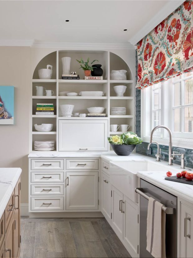 Bright, traditionally styled kitchen with extensive white built-in cabinetry, a marble-look benchtop, farmhouse-style sink, polished metal fixtures, and open display shelving filled with crockery. A large floral Roman blind adds colour beside the multi-pane windows, while grey timber-look flooring and subtle wall moulding complete the refined, lived-in space.