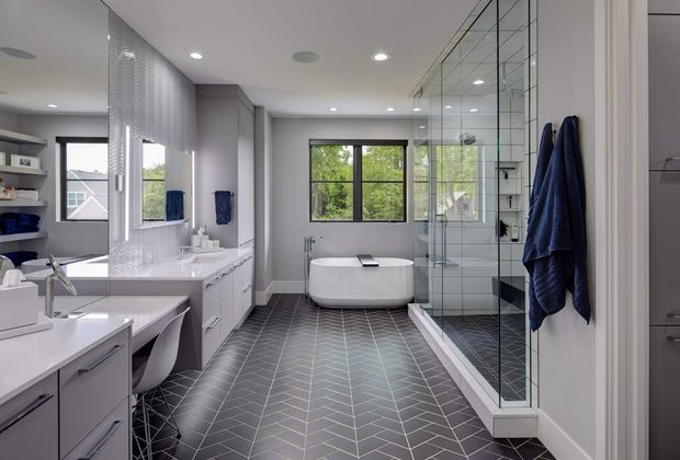 A spacious contemporary bathroom with a freestanding white bathtub positioned beneath a large window, a fully glazed walk-in shower, dual vanity with integrated storage, and a built-in makeup or dressing counter. Dark charcoal herringbone floor tiles contrast with pale grey walls and white fixtures, while navy towels provide a restrained accent. Mirrored and glass surfaces add brightness and visual depth.