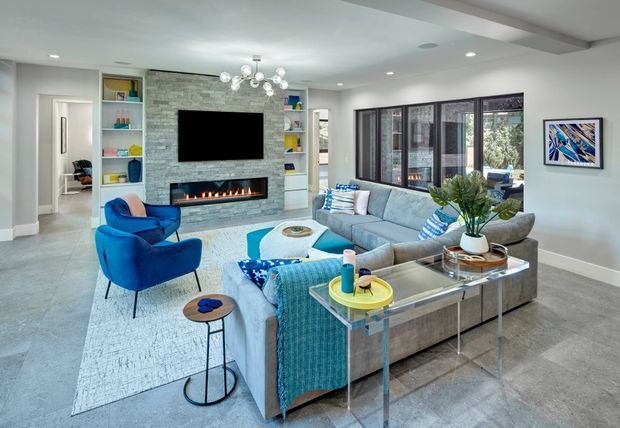 Bright, spacious contemporary living room with a large grey sectional sofa, vivid blue armchairs and textiles, a polished stone-look feature wall with built-in linear fireplace and television, open display shelving, extensive dark-framed glazing, and a sculptural chandelier. The room has a cool neutral palette with strong blue accents and a polished, welcoming atmosphere.