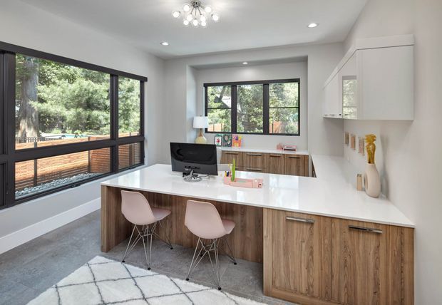 Bright contemporary home office with a large white island-style work surface and integrated timber cabinetry. Two blush-toned chairs sit at the island, while a desktop monitor, stationery and shelving create a functional workspace. The room has expansive black-framed windows overlooking greenery, recessed ceiling lights, a decorative modern pendant, white walls and grey carpet.