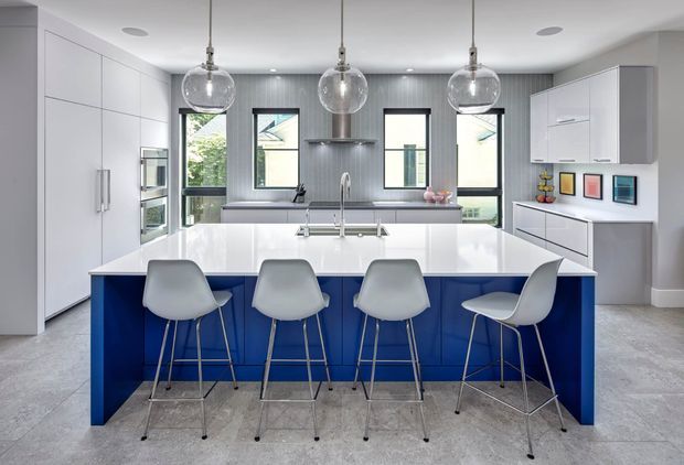 Bright contemporary kitchen with a large glossy white island featuring deep blue cabinetry and seating for four. The space includes full-height white cabinetry, integrated appliances, a grey vertically textured splashback, black-framed windows, pendant globe lights, and a pale grey tiled floor.