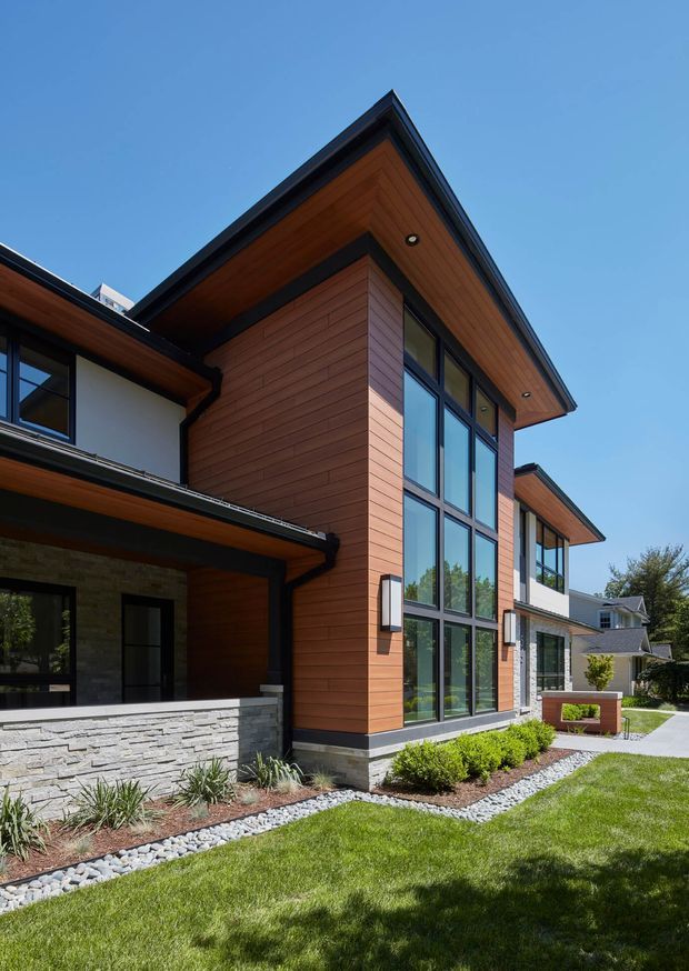 Exterior architectural photograph of a contemporary multi-level home viewed from the landscaped front garden. The house features warm horizontal timber cladding, dark-framed floor-to-ceiling glazing, light stacked-stone wall sections, broad timber-lined roof soffits, and crisp modern roof planes. A paved walkway, lawn, shrubs, and ornamental planting surround the facade.