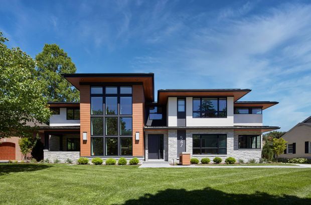 A wide exterior view of a large contemporary multi-level residence with strong horizontal roof planes, extensive dark-framed glazing, warm timber cladding, white rendered walls, and light grey stone veneer. The front facade opens onto a broad manicured lawn under a bright blue sky.