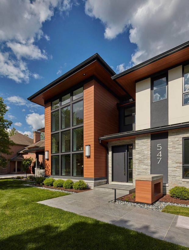 Street-facing exterior of a contemporary multi-storey residence with a prominent double-height glazed entry volume. The facade combines warm horizontal timber cladding, pale stone veneer, dark-framed windows and doors, and light rendered wall panels. A projecting flat roof with timber soffits shelters the entrance and upper-level forms. Concrete paths, clipped shrubs, lawn, and a built-in timber-and-stone mailbox or entry feature complete the frontage.
