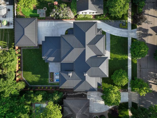 Aerial top-down view of a large residential property, dominated by a complex multi-section dark grey pitched and hipped roof. The house sits within a landscaped suburban lot with lawns, mature trees, garden beds, paved driveways and paths, and neighbouring homes visible around it.