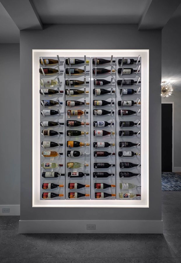 A dark, refined interior wall featuring a large illuminated built-in wine display. Multiple rows of wine bottles are held horizontally in a white-framed metal or glass rack, creating a dramatic feature installation. The surrounding walls and ceiling are charcoal grey, with a dark tiled floor and a decorative starburst wall light visible in the adjacent area.