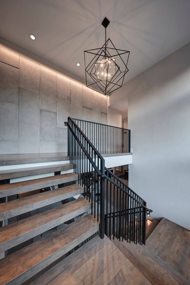 A contemporary interior stairway with open-riser timber stairs, black metal vertical balustrades, and a landing overlooking a lower level. Large grey concrete-look wall panels are softly illuminated by concealed linear lighting, while a sculptural black geometric pendant hangs from the ceiling. The space has a refined industrial character with warm timber flooring and dramatic stair geometry.