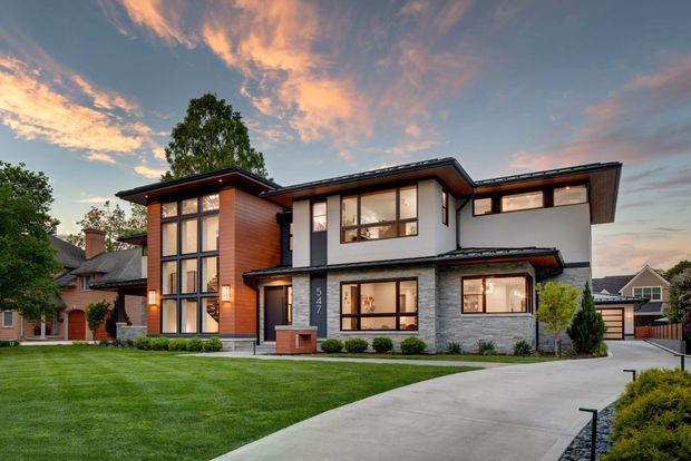 Exterior view of a large contemporary multi-storey residence at dusk, featuring broad floor-to-ceiling and horizontal windows, projecting low-pitched roof planes, dark trim, warm timber cladding, white rendered walls, and a grey stone-clad lower facade. A broad concrete driveway, manicured lawn, shrubs, and neighbouring homes surround the property.