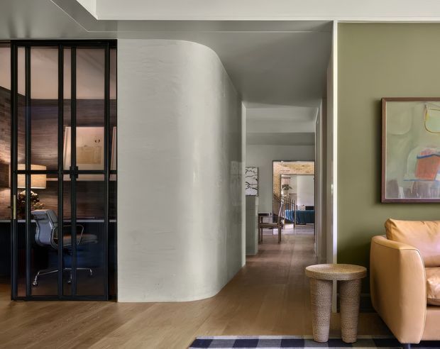 Interior view of a refined contemporary home hallway and living area. The composition features warm timber flooring, a sculptural curved plaster wall, a black-framed glazed partition enclosing a workspace or dining area, and a muted olive-green living-room wall with a tan leather sofa, woven side tables, and framed artwork. Additional rooms are visible along the central corridor.