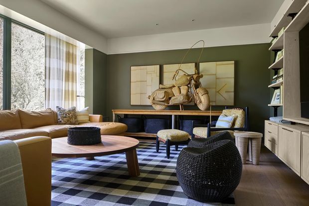 A stylish living room with a warm tan sectional sofa, low rounded timber coffee table, woven black ottoman and armchair, and a bold black-and-white checkered rug. The room features deep olive-green walls, white built-in shelving and cabinetry, large floor-to-ceiling windows with sheer curtains, and a sculptural artwork above a console. The overall aesthetic is richly layered, eclectic, and contemporary with natural woven textures and curved furniture forms.