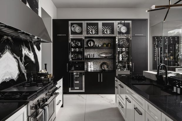 A sophisticated galley-style kitchen with a dramatic black-and-white palette. The room features dark cabinetry, white drawer fronts, a large professional gas range, black stone-look benchtops, a striking monochrome marble-pattern splashback, and a central wall of illuminated glass-front display shelving. Reflective surfaces and a sculptural linear pendant add a refined, high-contrast finish.