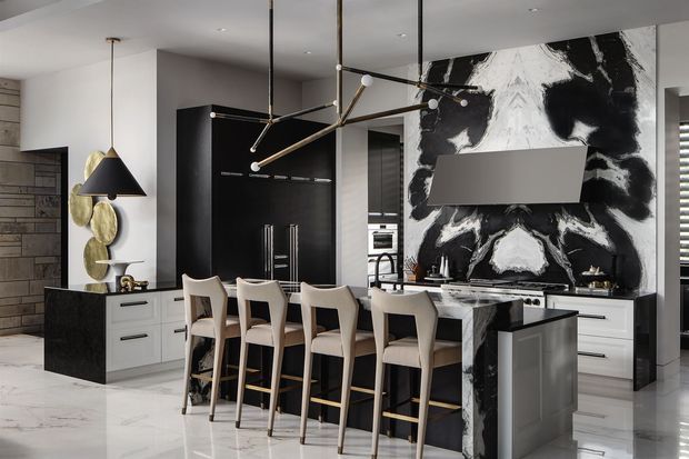A sophisticated open-plan kitchen dominated by a large island with seating for four, dark cabinetry, white drawers, and a dramatic black-and-white marble feature wall behind the cooktop. The space includes integrated appliances, pendant lighting, a sculptural linear chandelier, pale stone flooring, and a partial view of an adjacent dining or living area.
