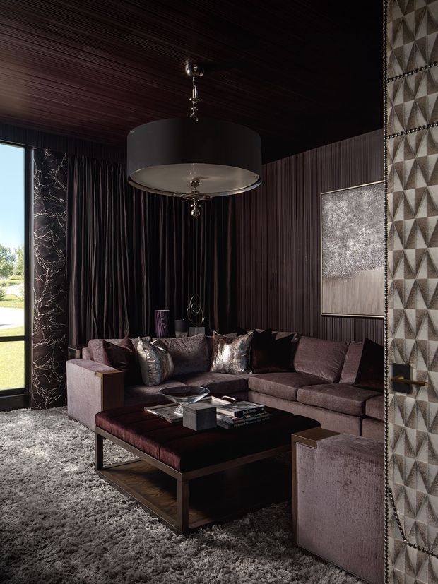 A dark, luxurious living room with a deep charcoal wood-slat ceiling and matching vertically textured wall treatment. A large sectional sofa, velvet armchairs, and a substantial dark coffee table sit on a plush grey rug. The space features metallic cushions, a sculptural pendant light, a marble-look column, framed artwork, and a geometric upholstered wall or door panel, creating a dramatic, layered interior.