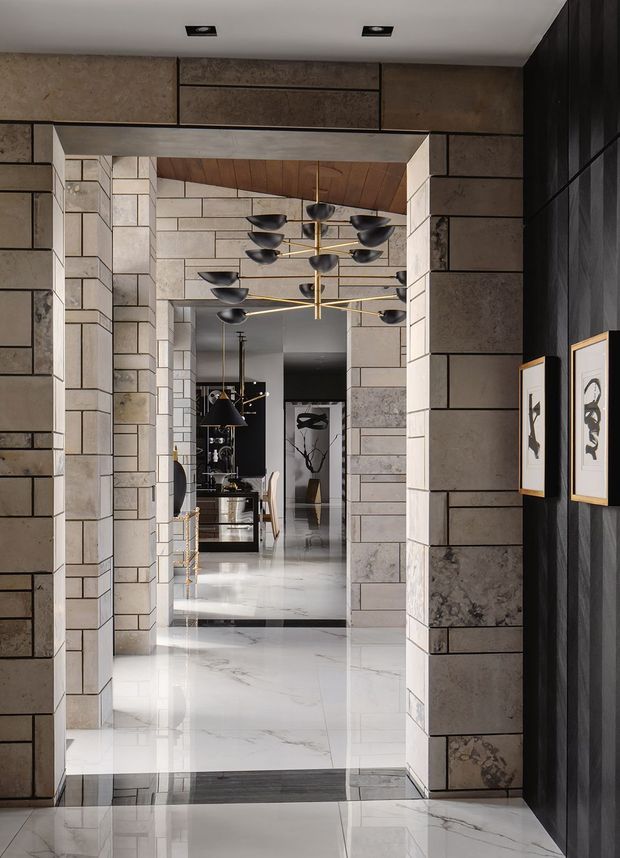 A refined interior hallway framed by substantial rectangular stone-clad wall panels and door surrounds. The polished white marble-like floor reflects a sculptural brass-and-black multi-arm chandelier, while a timber-lined ceiling recess adds warmth. The corridor opens toward a dining or living area with dark cabinetry, pendant lighting, seating, and artwork. Dark vertical wall panelling and framed monochrome art create a dramatic, high-contrast finish.