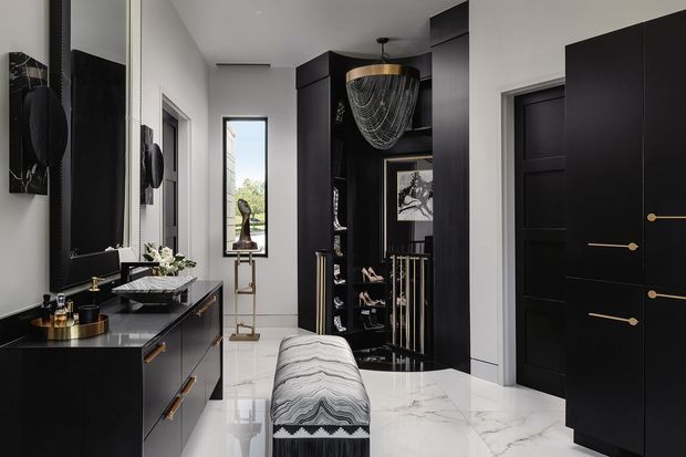 A sophisticated residential entry hall with a long black built-in console and mirror on the left, a central upholstered bench, black paneled doors and tall dark cabinetry, and a sculptural pendant light above an open display and coat-storage area. The space features glossy white marble-look flooring with grey veining, crisp white walls, and dramatic black joinery with brass hardware.