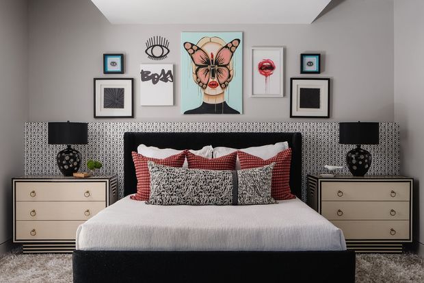 A symmetrical, boldly styled bedroom with a low black upholstered bed, layered white and patterned bedding, red accent cushions, matching cream bedside chests, black lamps, and a graphic black-and-white patterned feature wall. A curated gallery of colorful and monochrome artwork hangs above the bed. The room has a dramatic, eclectic and refined feel, with a recessed ceiling detail and plush rug.