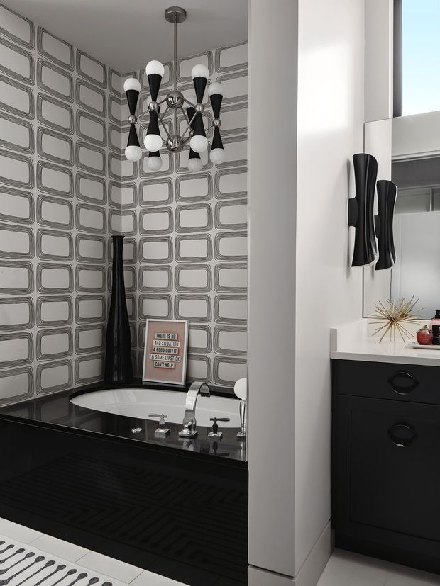A dramatic black-and-white bathroom featuring a freestanding bathtub set into a dark built-in surround, chrome tapware, and a bold geometric rectangular-patterned wall finish. A sculptural black-and-white chandelier hangs above the bath, while a black vanity with curved drawer pulls and matching wall sconces is visible to the right. The space has a high-contrast, glamorous retro-modern character.