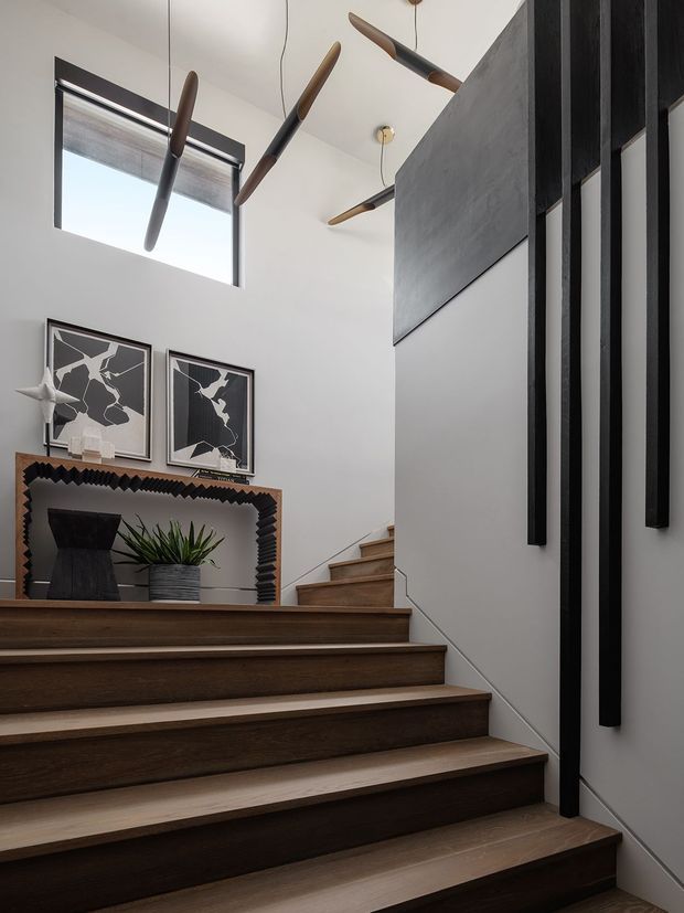 Interior view of a modern stairway with broad dark timber stair treads, white walls, and a black vertical-slat feature wall. The stair landing includes a timber console with a dark textured surround, framed monochrome artwork, a potted plant, and sculptural pendant lights with timber accents. A high window brings in natural light.