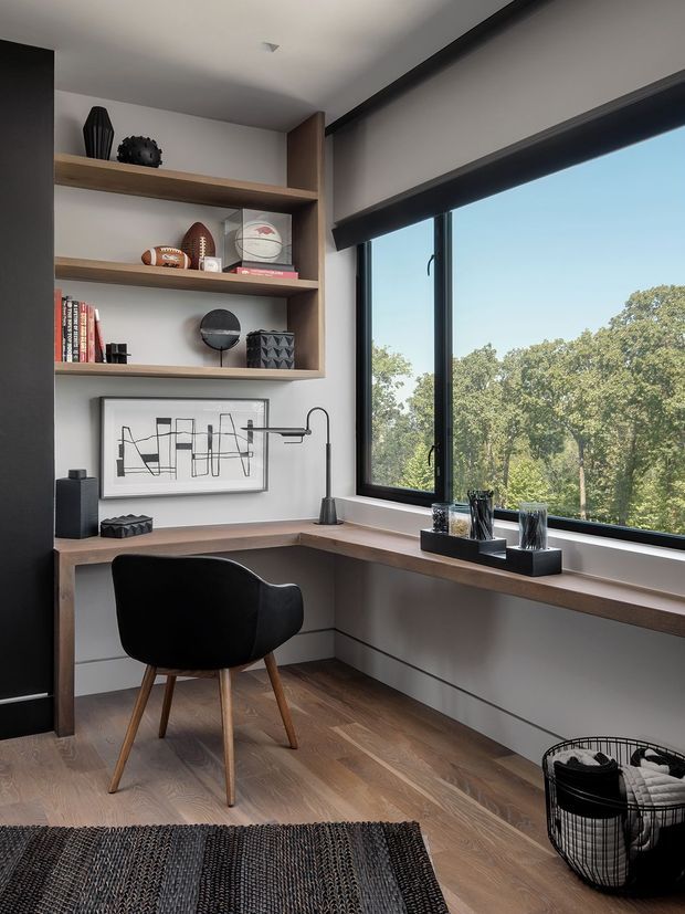 A compact, contemporary home office nook with a built-in L-shaped timber desk beneath a large black-framed window. Open timber shelving displays books, sports memorabilia and sculptural objects, while a black upholstered chair, task lamp, artwork and dark woven rug complete the workspace. The room has light neutral walls, timber flooring, dark joinery and expansive views of trees through the window.
