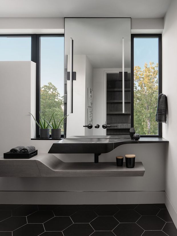 A contemporary bathroom vanity area with a large frameless mirrored cabinet, wall-mounted dark basin and integrated countertop. Black hexagonal floor tiles, dark timber shelving visible in the mirror, slim black-framed windows, and minimalist monochromatic finishes create a refined, high-contrast interior. Potted plants and folded towels add subtle natural and functional details.