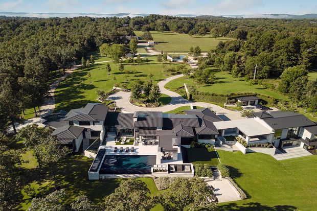Aerial view of a large contemporary residential estate set within expansive landscaped grounds and mature woodland. The multi-wing home has dark pitched and hipped metal roofs, light exterior walls, extensive glazing, multiple terraces, and a large rectangular swimming pool with surrounding paved outdoor areas. Curving driveways and paths connect the house to lawns, gardens, and additional structures, with rolling wooded terrain visible beyond.