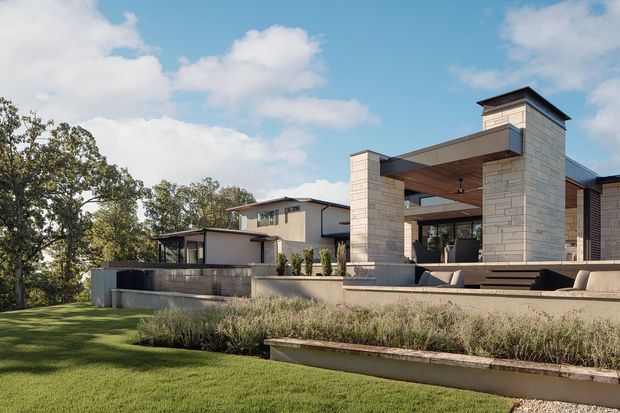 Exterior view of a large contemporary residence set within a landscaped garden. The composition highlights a covered outdoor entertaining pavilion with substantial pale masonry piers, a broad flat roof, timber-toned soffit, integrated ceiling fan and steps leading to raised terraces. A smaller light-coloured house volume sits behind it, with mature trees and lawn surrounding the property.