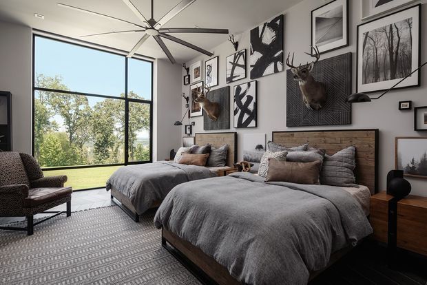 Spacious contemporary bedroom with two large beds, grey bedding, dark timber bed frames and integrated wood headboards. A dramatic gallery wall of black-and-white artwork and mounted deer heads forms the main feature, while floor-to-ceiling glazing overlooks a green landscape. A large ceiling fan, armchair, bedside tables and textured rug complete the room.