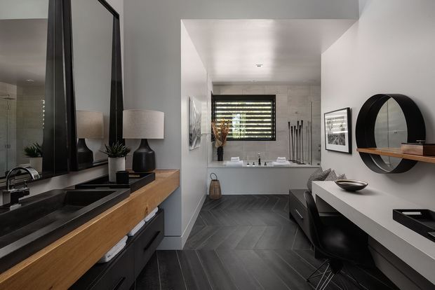 A contemporary bathroom with a long, open vanity and integrated black basins in the foreground, warm timber drawer fronts, matte black tapware, and large black-framed mirrored panels. The room extends toward a freestanding bathtub beneath a horizontal louvred window, with dark geometric floor tiles, pale walls, a wall-mounted desk or dressing counter, black seating, framed artwork, and a circular black wall mirror with a timber shelf. The palette is refined and high-contrast, combining charcoal, white, and timber finishes.