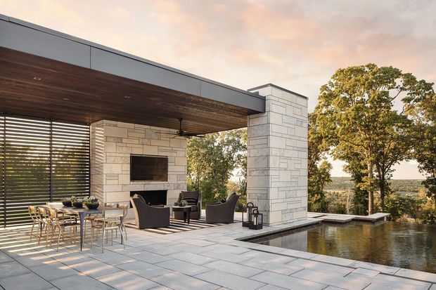 A refined modern outdoor entertaining terrace beside an infinity-edge pool, with a covered dining and lounge area, large-format pale stone paving, a dark timber-lined ceiling, stone-clad structural columns and walls, horizontal timber screening, and an outdoor fireplace with wall-mounted television. The terrace overlooks a wooded landscape in warm late-day light.
