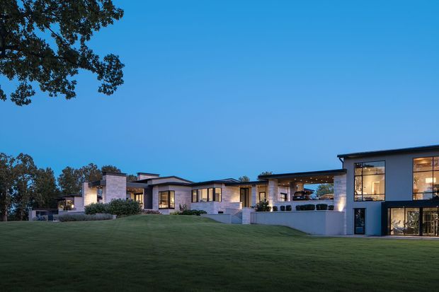 Wide dusk exterior view of a sprawling contemporary residence set behind an expansive manicured lawn. The home has a low, horizontally composed form with flat and shallow-pitched roof sections, pale masonry or rendered wall surfaces, dark window and door frames, extensive glazing, and warm interior lighting visible through the windows. Mature trees frame the property.