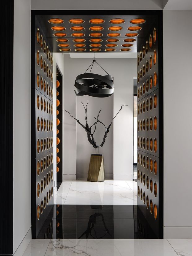 A dramatic, highly refined interior passage or entry feature framed by dark metallic perforated wall and ceiling panels with repeated warm copper-toned circular insets. A sculptural black ring pendant hangs centrally above a tall geometric metallic vessel containing branching dark sculpture. Glossy white marble flooring with a black reflective inset creates strong symmetry and reflection, while concealed lighting and monochrome finishes produce a luxurious, gallery-like atmosphere.