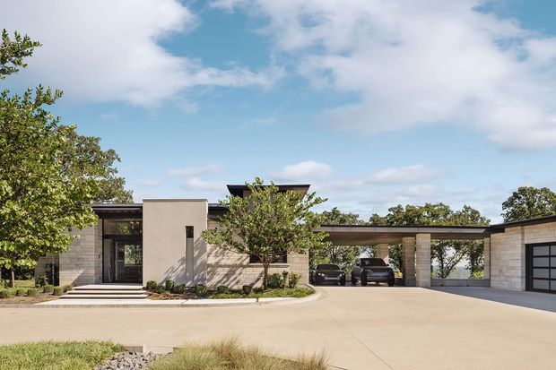 Contemporary single-storey residential exterior with a low, elongated roofline, pale masonry walls, smooth rendered feature volumes, dark-framed glazing, and a covered carport/garage wing. A broad concrete driveway and simple landscaped planting are visible in the foreground.