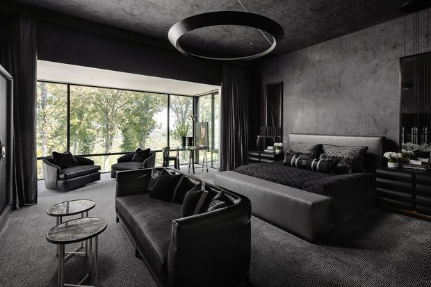 A sophisticated, dark-toned living room with a large sectional sofa, black leather lounge chairs, nested round coffee tables, textured grey carpet, dark curtains, and a dramatic circular pendant light. Floor-to-ceiling corner glazing opens the room to leafy views, while textured charcoal walls and dark built-in joinery create a moody, luxurious atmosphere.