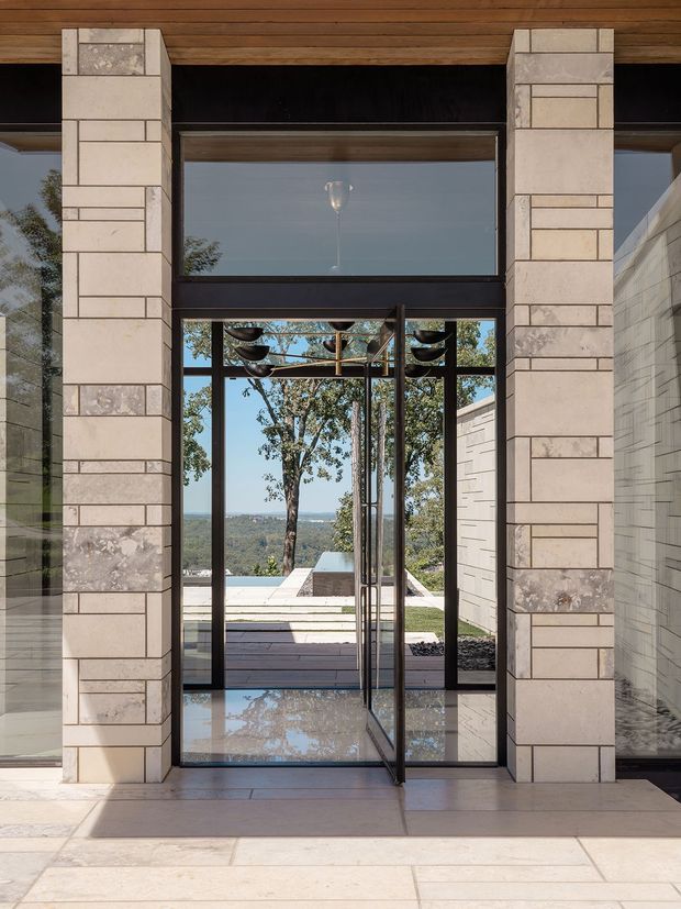 A dramatic residential entryway viewed from inside, framed by substantial pale stone-clad piers and dark metal-framed floor-to-ceiling glazing. A glass pivot door opens toward a terrace and distant wooded landscape, while a timber-lined ceiling and reflective polished stone floor add warmth and visual depth. A sculptural pendant is visible through the glazed entry.