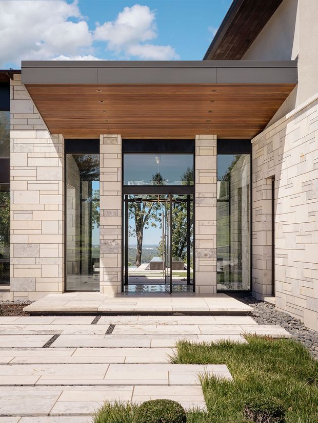 A contemporary residential entrance viewed from the landscaped approach. Pale rectangular stone cladding forms the walls and columns, with a warm timber-lined cantilevered canopy above a tall black-framed glazed entry. Broad pale stone paving leads to the doorway, bordered by dark gravel and clipped grasses. The glazing reflects trees and reveals a glimpse of the interior and distant landscape beyond.