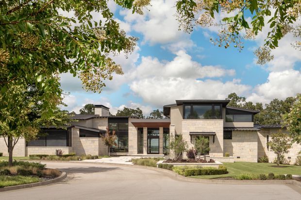 Wide exterior view of a low, sprawling contemporary residence set behind a curved driveway and landscaped frontage. The home uses pale masonry block walls, dark metal-framed glazing, strong horizontal rooflines, recessed entryways, and a taller central glazed volume. Mature trees, clipped planting beds, lawn, and ornamental grasses frame the property.