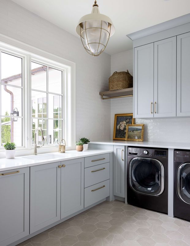 Bright, refined laundry room with pale grey-blue shaker cabinetry, white benchtops, a large divided-pane window, integrated front-loading black washer and dryer, and a white tiled backsplash. Brass hardware, a woven basket, framed artwork, and a sculptural pendant add warm decorative detail. The floor is finished in large pale hexagonal tiles.