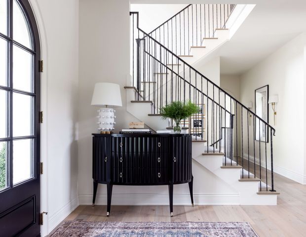 Bright, refined residential entry hall featuring a black arched glazed entry door, a dark fluted sideboard with lamp and greenery, pale timber flooring, and a staircase with slender black metal balustrades. White walls, crisp trim, and abundant natural light create a polished transitional interior.