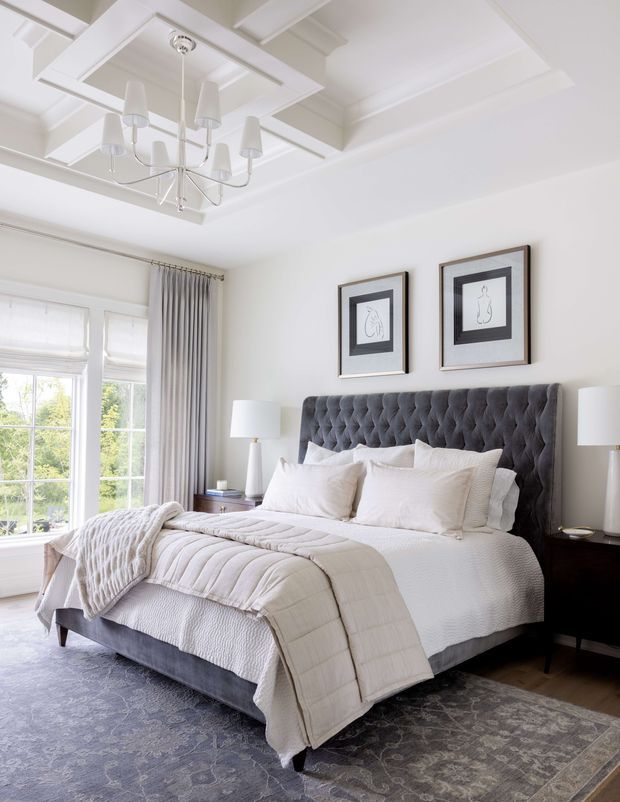 Bright, refined bedroom with a large tufted charcoal-grey upholstered bed, layered white bedding, dark bedside tables, matching lamps, framed artwork, full-height curtains, and a patterned grey area rug. The room features a white coffered ceiling with a contemporary chandelier and tall windows bringing in abundant daylight.