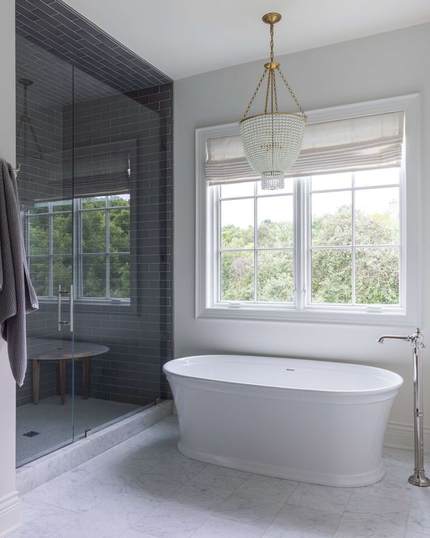 Elegant, bright bathroom featuring a freestanding white bathtub beside a large multi-pane window, a glass-enclosed walk-in shower with dark charcoal subway tile, pale marble-look floor tile, grey painted walls, and a decorative crystal chandelier with brass-toned fittings.
