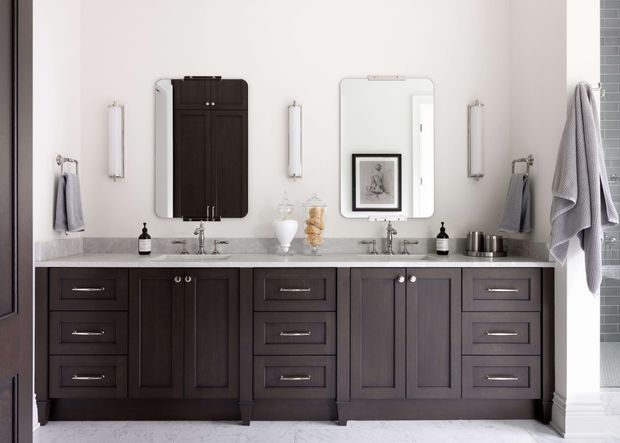 A refined double-vanity bathroom with dark timber-look cabinetry, pale stone countertop, paired undermount sinks and polished metal faucets. Two framed mirrors, slim vertical wall lights, and symmetrical drawer and cupboard storage create a balanced traditional design. Towels and toiletries are visible, with a glimpse of a tiled shower area at right.