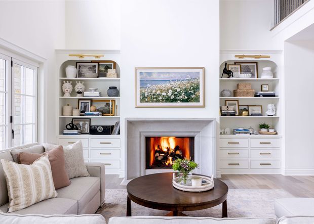 Bright, refined living room centered around a traditional fireplace with a firebox and pale stone surround. Symmetrical built-in white shelving and drawer cabinetry flank the fireplace, displaying books and decor. A large framed artwork hangs above the mantel, while upholstered sofas, patterned cushions, a dark round coffee table, and a pale area rug create a comfortable seating arrangement. French-style glazed doors are visible at left, and an upper-level balustrade is visible at right, suggesting a high or partially double-height space.