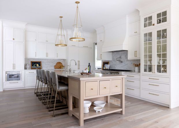 Bright, spacious white kitchen with extensive built-in cabinetry, a large central island with seating for four, a farmhouse-style sink, marble-look backsplash, integrated appliances, glass-front display storage, and three elegant pendant lights. Light timber flooring and warm taupe island cabinetry add contrast to the predominantly white scheme.